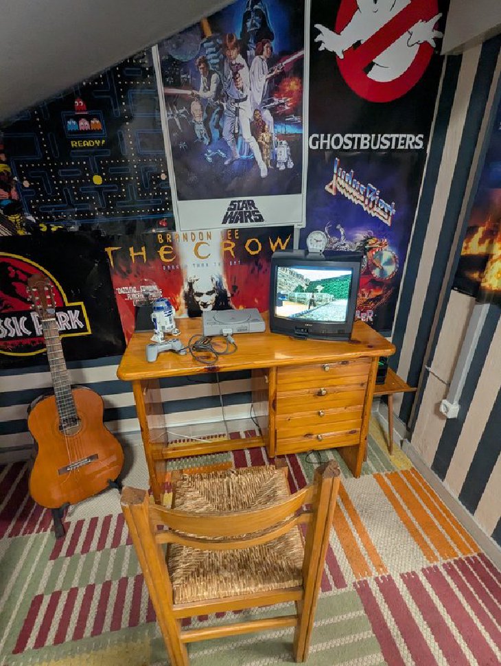 A recreated 1990s bedroom with a PlayStation and a small CRT TV running Tomb Raider II on a pine desk, surrounded by Star Wars, Ghostbusters and The Crow posters